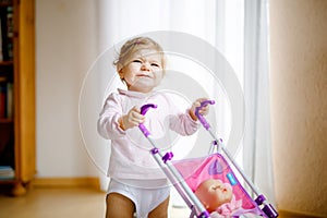 Cute adorable baby girl making first steps with doll carriage. Beautiful toddler child pushing stroller with toy at home