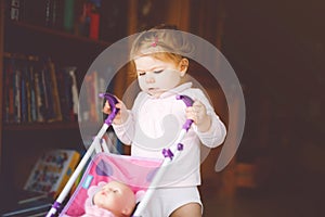Cute adorable baby girl making first steps with doll carriage. Beautiful toddler child pushing stroller with toy at home