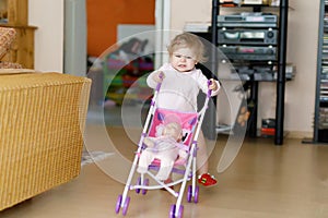 Cute adorable baby girl making first steps with doll carriage.