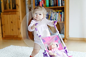 Cute adorable baby girl making first steps with doll carriage.