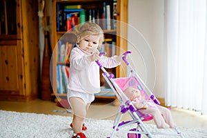 Cute adorable baby girl making first steps with doll carriage. Beautiful toddler child pushing stroller with toy at home