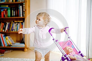 Cute adorable baby girl making first steps with doll carriage. Beautiful toddler child pushing stroller with toy at home