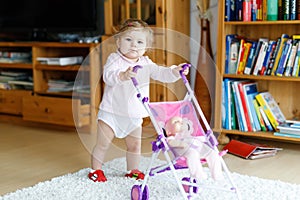 Cute adorable baby girl making first steps with doll carriage.