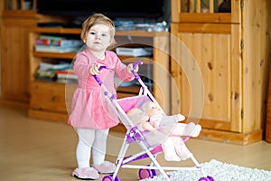 Cute adorable baby girl making first steps with doll carriage.