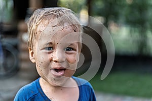 Cute adorable blond caucasian little happy toddler boy portrait with messy mus spots on face after playing watering