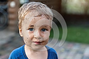 Cute adorable blond caucasian little happy toddler boy portrait with messy mus spots on face after playing watering