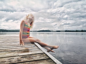 Cute adorable blonde Caucasian girl sitting on wooden dock by lake and looking up in the sky. Dreaming relaxing cute child kid by