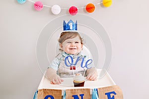 Cute adorable Caucasian baby boy in blue crown celebrating first birthday at home. Child kid toddler sitting in high chair eating