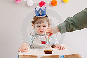 Cute adorable Caucasian baby boy in blue crown celebrating first birthday at home. Child kid toddler sitting in high chair looking