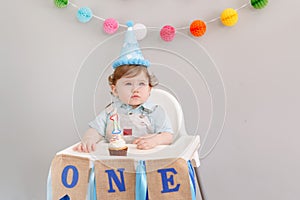 Cute adorable Caucasian baby boy in blue hat celebrating his first birthday at home. Child kid toddler sitting in a high chair