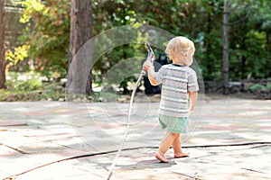 Cute adorable caucasian blond barefeet boy walking at home backyard holding hose pipe for watering garden. Child little