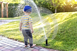 Cute adorable caucasian blond barefeet toddler boy in cap walking at home backyard near sprinkler automatic watering
