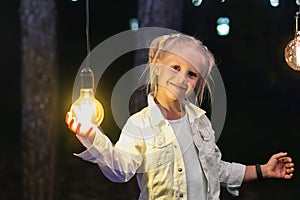 Cute adorable caucasian blond girl portrait smiling and holding in hand one of hanged edison light bulb at forest outdoor. Right