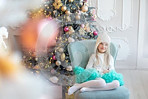 Cute adorable caucasian blond little kid girl sitting in chair at home near christmas tree and dreaming of christmas