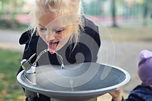 Cute adorable caucasian blond little thirsty school girl drinking water from public potable fountain faucet in city park