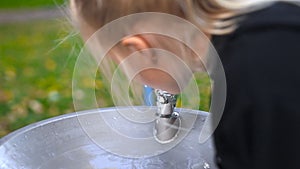 Cute adorable caucasian litte blond girl drinking water from faucet with drinking water fountain at city park near
