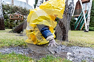 Cute adorable caucasian toddler boy in bright yellow raincoat and wellies playing alone at dirt muddy puddle during cold