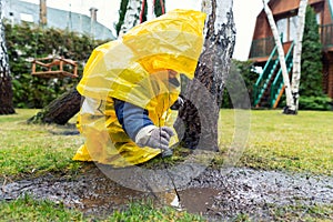 Cute adorable caucasian toddler boy in bright yellow raincoat and wellies playing alone at dirt muddy puddle during cold spring