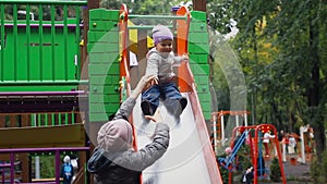 Cute adorable caucasian toddler boy having fun sliding down wet metal slide at playground in city park after rain in