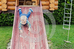 Cute adorable caucasian toddler boy having fun sliding down wooden slide at eco-friendly natural playground at backyard in autumn