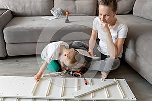 Cute adorable caucasian toddler boy kid sit on floor and help mom assembling furniture shelf with power screwdriver tool