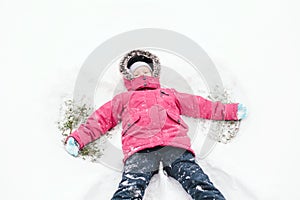 Cute adorable funny Caucasian girl child in warm clothes pink jacket making snow angel. Kid lying on ground during a cold winter