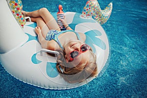 Cute adorable girl in sunglasses with drink lying on inflatable ring unicorn. Kid child enjoying having fun in swimming pool.