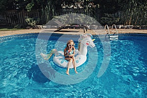 Cute adorable girl in sunglasses with drink lying on inflatable ring unicorn. Kid child enjoying having fun in swimming pool.