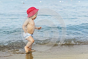 Cute adorable little blond caucasian serious pensive toddler boy in red panama walking through blue clear water waves of