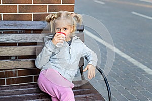 Cute adorable little caucasian blond kid girl enjoy sitting om wooden bench and drink hot chocolate drink with paper cup. Beatiful