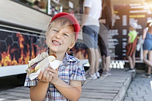 Cute adorable little happy smiling boy kid enjoy eating hot dog sausage in bread near street cafe stall outdoors. Child