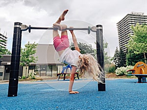 Cute adorable little preschool blonde Caucasian girl hanging upside down on pullup bar on playground.