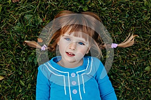 cute adorable little red-haired Caucasian girl child in blue dress lying in field meadow park outside