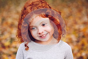 Cute adorable smiling little red-haired Caucasian girl child standing in autumn fall park outside, looking away
