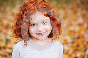 Cute adorable smiling little red-haired Caucasian girl child standing in autumn fall park outside, looking away