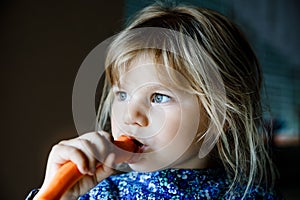 Cute adorable toddler girl holding and biting into fresh carrot. Beatuiful child having healthy snack. Smiling happy kid