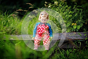 Cute adorable toddler girl sitting on wooden bridge and throwing small stones into a creek. Funny baby having fun with