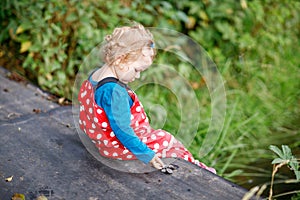 Cute adorable toddler girl sitting on wooden bridge and throwing small stones into a creek. Funny baby having fun with