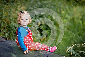 Cute adorable toddler girl sitting on wooden bridge and throwing small stones into a creek. Funny baby having fun with