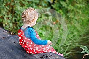 Cute adorable toddler girl sitting on wooden bridge and throwing small stones into a creek. Funny baby having fun with