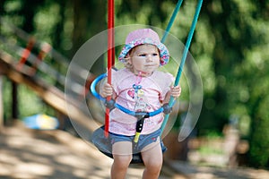 Cute adorable toddler girl swinging on outdoor playground. Happy smiling baby child sitting in chain swing. Active baby