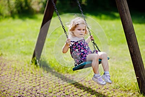 Cute adorable toddler girl swinging on outdoor playground. Happy smiling baby child sitting in chain swing. Active baby