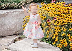 Cute adorable white Caucasian baby girl child in white dress standing among yellow flowers outside in garden park