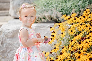 Cute adorable white Caucasian baby girl child in white dress standing among yellow flowers outside in garden park looking in