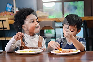 Cute African American boy with curly and adorable Asian kid eating meal at the table indoor, happy children having food in a