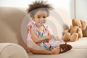 Cute African American child imagining herself as doctor while playing with stethoscope and doll on couch