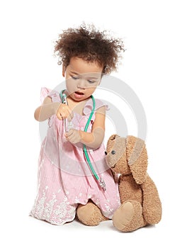 Cute African American child imagining herself as doctor while playing with stethoscope and toy bunny on white