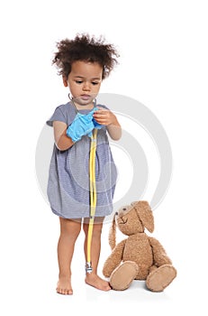 Cute African American child imagining herself as doctor while playing with stethoscope and toy bunny on white