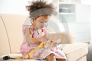 Cute African American child imagining herself as doctor while playing with stethoscope and toy on couch