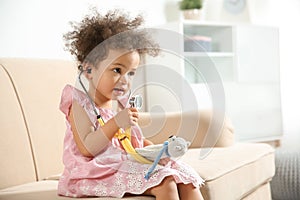 Cute African American child imagining herself as doctor while playing with stethoscope and toy on couch